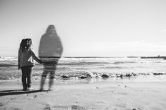 A young girl stands before the sea shore holding hands with the transparent, ghostly figure a deceased person who is dear to her. (a memory)
