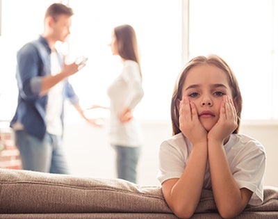 A young girl looks distraught while her parents argue in the background.