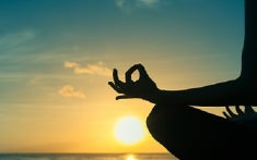 A silhouetteof a woman sitting cross legged with her wrists resting on her knees and fingers in the om position in front of a sunset.