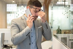 A business man pinches his nose while on the phone looking stressed.