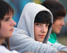Young man perhaps high school or college age with a heather gray sweatshirt hoodie  pulled over his head looking at you.