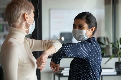 Two women bump elbows while wearing masks.