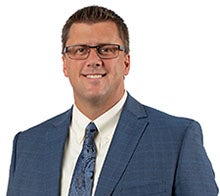 A man with short hair and glasses wearing a cream shirt with a blue suit and tie.