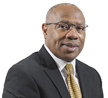 African American man in dark suit with gold tie and glasses.