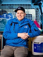 Matt Amendt sitting in the back of a work van smiling at the camera wearing a bright blue work jacket and baseball cap.