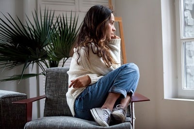 A young woman with brown wavy hair who is sitting on a chair with her legs drawn up to her chest while sadly gazing out a window.
