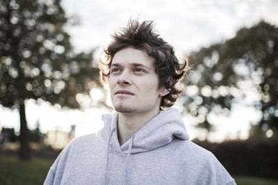 A pale-faced young man with messy hair wearing a grey sweatshirt looking up and away toward something hopeful.