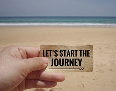 Someone's hand holding a paper that says Let's Start the Journey with the beach in the background.
