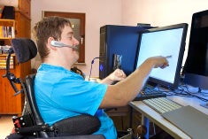 A younger man sitting in a wheelchair at his desk pointing at a computer screen.