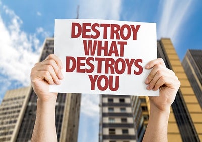 Person holding a sign in front of a tall building that reads Destroy What Destroys You.