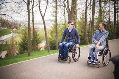 Two young men in wheelchairs rolling through park while laughing and talking.