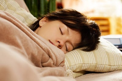 A young woman with brown hair is under a blanket with her head on a pillow asleep.