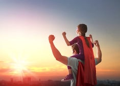 A father and son with superhero capes posing with arms up and looking toward the bright sun.