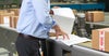 Close-up of man's torso and hands as he examines paperwork on a conveyor belt alongside cardboard boxes
