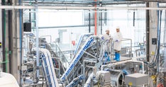 Two workers in white lab coats, hard hats, gloves, and blue rubber boots stand on a metal platform inside a bright, industrial food processing facility, overseeing stainless steel conveyor systems, pipes, and automated machinery.