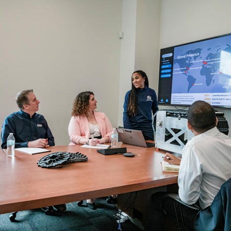 People in a boardroom watching a woman present on a television screen