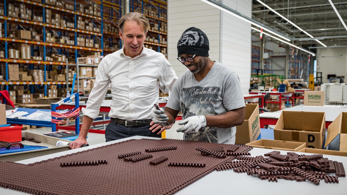Two men are smiling while one assembles a brown modular plastic conveyor belt inside a well-organized industrial warehouse.