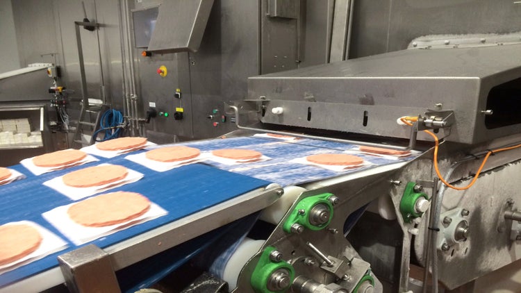 Raw burger patties on tissue paper entering freezer spiral conveyor