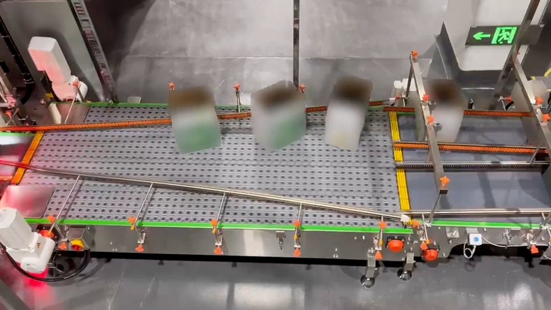 Un nastro trasportatore che movimenta confezioni, circondato da diversi componenti meccanici e barriere di sicurezza.