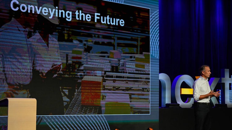 Jeroen Neuhof, camicia bianca e pantaloni scuri, parla sul palco del NEXT 2024. Sullo sfondo, appare un grande schermo con la frase "Conveying the Future" (Portiamo avanti il futuro).