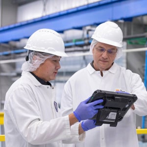 Zwei Männer in weißen Laborkitteln und mit Helmen, die in einer Fabrik auf ein elektronisches Tablet blicken
