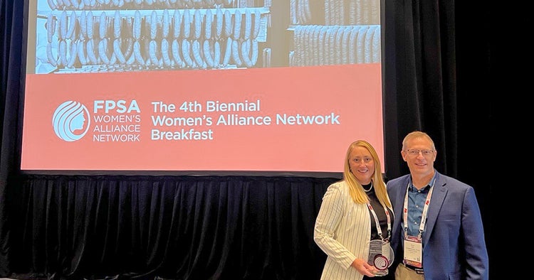 Eine Frau und ein Mann lächeln vor einem roten Hintergrund mit der Aufschrift „FPSA Women's Alliance Network: The 4th Biennial Women's Alliance Network Breakfast“. Die Frau, Nicole Cammarata, hält die Plakette der ihr verliehenen FPSA Red Circle Honor in Händen.