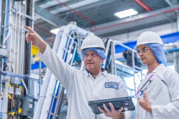 Two men in protective gear, including lab coats, helmets, and hairnets. One of the men is looking at and pointing to something out of frame, the other wears glasses and is looking at the same thing. Background is factory.