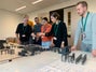 Eine Gruppe von Personen (zwei Frauen und drei Männer sichtbar) steht an einem Tisch und arbeitet in einem Workshop zur Förderbandkonstruktion unter Verwendung von Bauplänen und Maßstabsmodellen von Förderern zusammen.