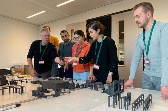 A group of people (two women and three men visible) standing over a table, collaborating in a conveyor design workshop using blueprints and scale models of conveyors.
