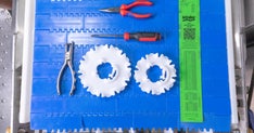 Top-down view of a blue modular plastic conveyor belt with various tools neatly arranged on it. Items include two white plastic sprockets, a pair of pliers, needle-nose pliers with red handles, a screwdriver, and a bright green Intralox belt replacement ruler.