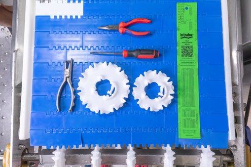Top-down view of a blue modular plastic conveyor belt with various tools neatly arranged on it. Items include two white plastic sprockets, a pair of pliers, needle-nose pliers with red handles, a screwdriver, and a bright green Intralox belt replacement ruler.