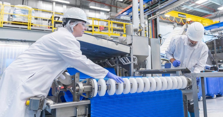 Dois funcionários, vestindo jalecos brancos e equipamento de proteção, examinando uma esteira transportadora FoodSafe azul e engrenagens de roda em uma fábrica