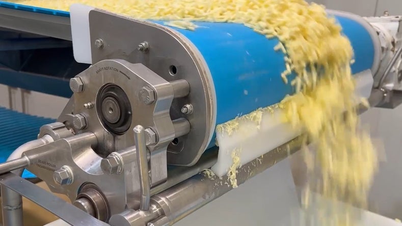 Close-up of shredded cheese moving on a blue conveyor belt in a stainless steel food processing machine, falling off the end in a steady stream.