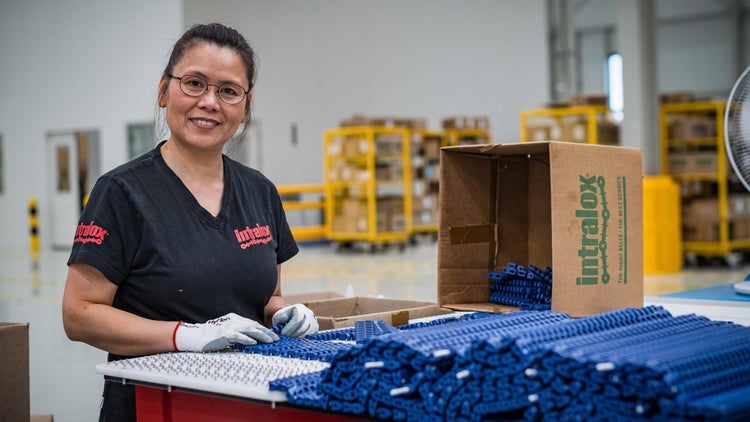 Una mujer con una camiseta negra con el logotipo de Intralox monta una banda transportadora de plástico azul en una estación de trabajo. A su lado hay una caja de cartón abierta con el logotipo de Intralox.