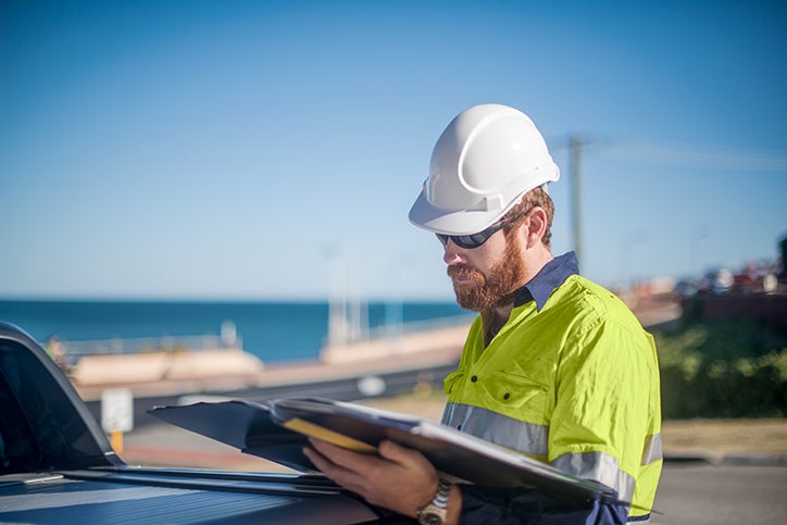 Construction worker with beard wearing hat and sunglasses looking at book.