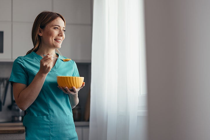 A nurse in green scrubs eating breakfast and looking out the window