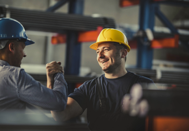 Two industrial workers in a manufacturing plant, one wearing a blue hard hat and the other in a yellow hard hat, collaborating over machinery