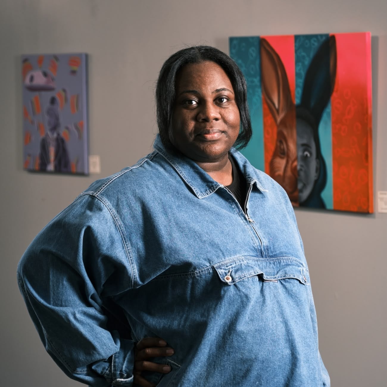 Black-haired woman wearing a black undershirt and a denim jacket stands in an art gallery looking at the camera with a neutral expression