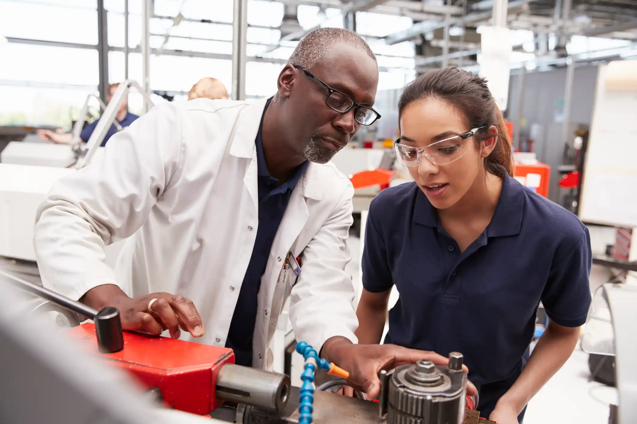 Engineering apprentice in a lab wearing protective eyewear next to the tutor in a lab coat