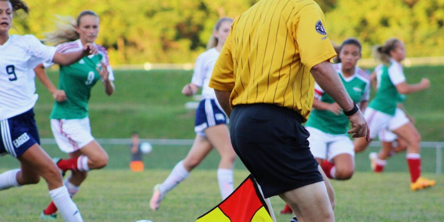 Young women playing soccer - green and white uniforms versus white and blue uniforms, referee in foreground wearing yellow shirt and black shorts holding a red and yellow flag
