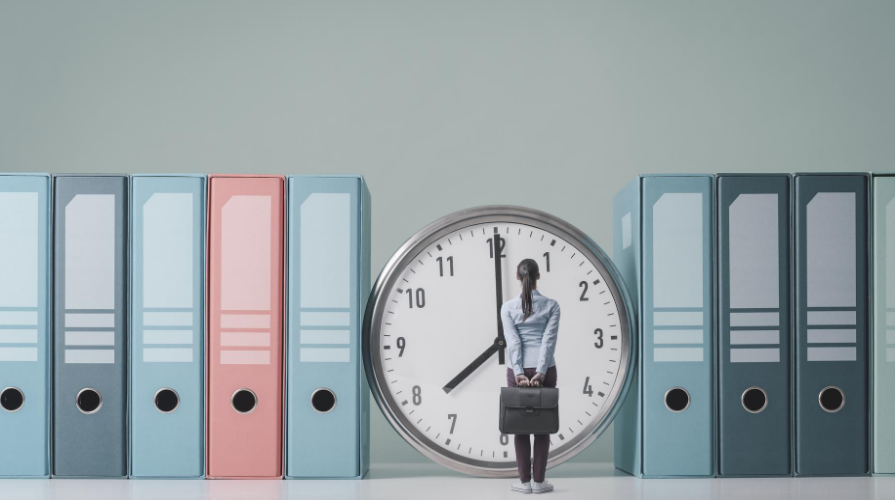 photo of a miniature sized woman standing, facing a clock, holding a briefcase behind her back, different shades of blue and pink binders line the wall on both sides of the clock 