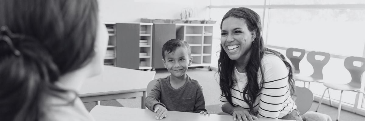 Black and white photo of a parent and young boy student meeting with a teacher in a classroom. 