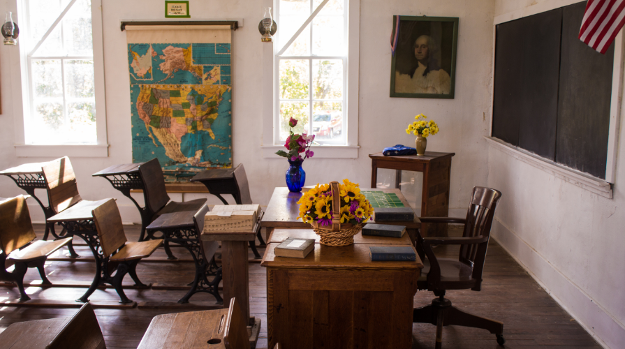 photo of an empty classroom filled with wooden desks, map of the United States, and a basket of sunflowers on the teacher's desk