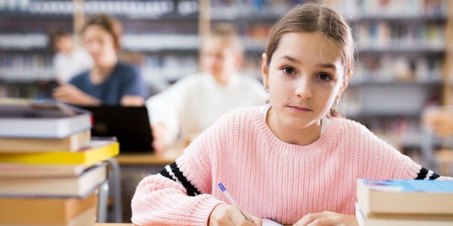 Young girl student in library sitting at table with pen and paper, staring into camera looking serious