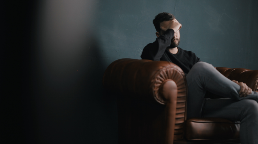 photo of a sad, stressed man sitting on a brown leather couch with his head resting on his right hand