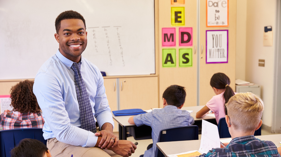 photo of a black male teacher smiling and sitting on a desk in a classroom