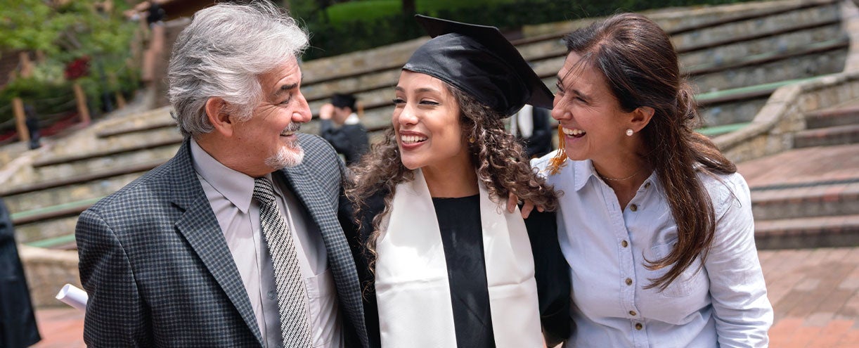 An image of a father and mother hug their daughter who has just graduated and is wearing her cap and gown.