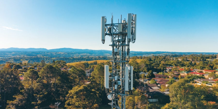 photo of the top of a cell phone tower on a sunny day with clear blue skies, overlooking a neighborhood tucked beneath tall green trees