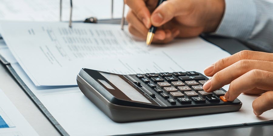 photo of a person working on a budget with left hand typing on a calculator and right hand holding a pen over a document