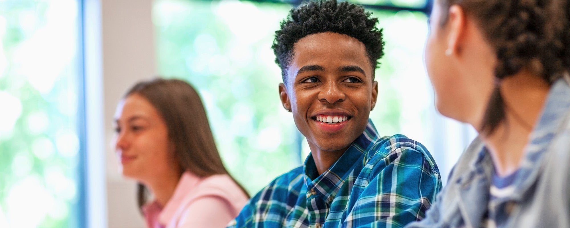 photo of a student in a classroom, wearing a plaid blue shirt and smiling at a girl sitting next to him